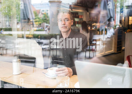 Man sitting in a cafe behind windowpane, drinking coffee with laptop Stock Photo