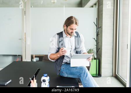 Young businessman sitting on table in office with tablet and cup of coffee Stock Photo
