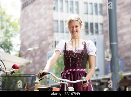 Portrait of smiling blond woman with bicycle wearing dirndl in the city, Freiburg, Germany Stock Photo