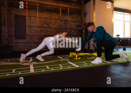Young woman exercising in fitness gym with personal trainer pushing weight sled Stock Photo