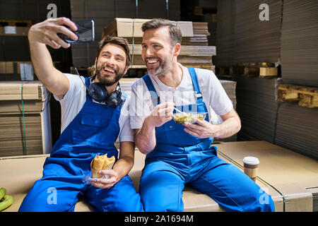 Two happy workers having lunch break and taking a selfie in factory Stock Photo
