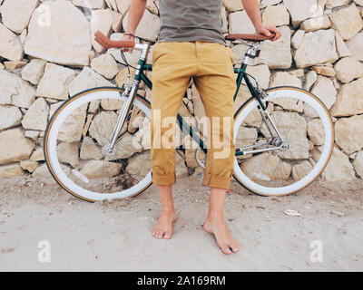 Barefoot man with Fixie bike in front of natural stone wall on the beach, partial view