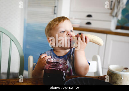 Cute little boy eating banana with blueberry jam in the kitchen Stock Photo