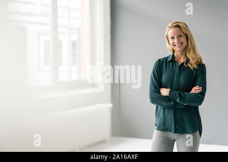Successful, blond businesswoman standing in bright room, with arms crossed Stock Photo