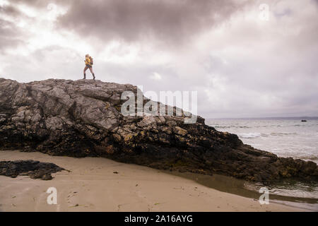 Woman wearing yellow rain jacket standing at rocky beach Stock Photo