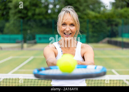 Portrait of laughing mature woman holding a tennis racket with a ball at tennis club Stock Photo