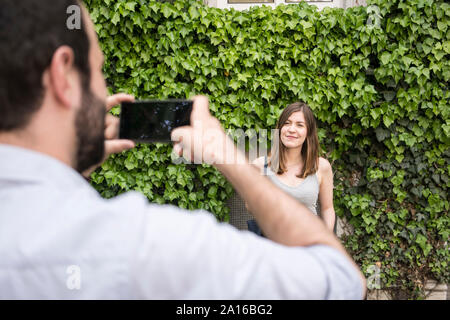 Man taking cell phone picture of young woman at a hedge Stock Photo