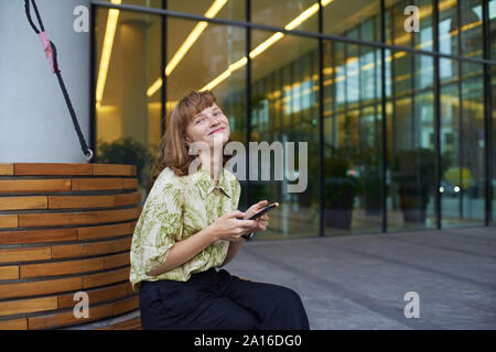 Young redheaded female model with freckles sitting at the wooden bench near modern business center and holding her smartphone smiling Stock Photo