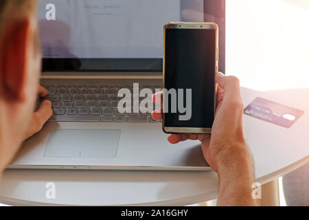 Smartphone with empty black screen in the hand of man using laptop and going to make payment with phone. Stock Photo