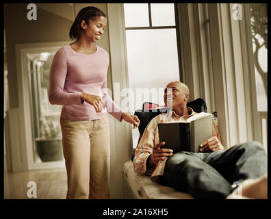 Mid-adult man lying on a window seat reading while his wife stands beside him in their home. Stock Photo