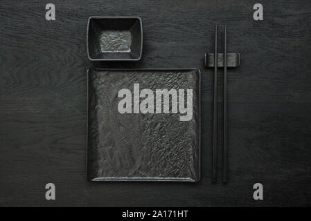 Black chopsticks on chopsticks rest, empty square slate plate and bowl for soy sauce on black wooden table. Flat lay. Mock-up. Asian food concept. Top Stock Photo