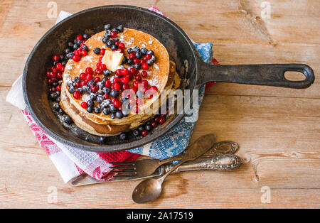 Stack of homemade oat pancakes with powered sugar, blueberries and cowberries in cast iron pan on rustic country table with old silver tableware. Soft Stock Photo