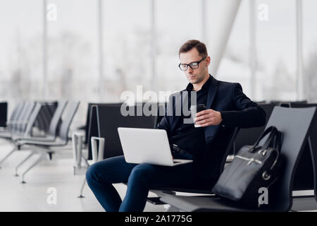 Handsome young businessman working on a laptop and drinking a coffee during an expectation of a flight. Stock Photo