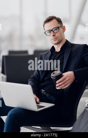 Handsome young businessman working on a laptop and drinking a coffee during an expectation of a flight. Stock Photo