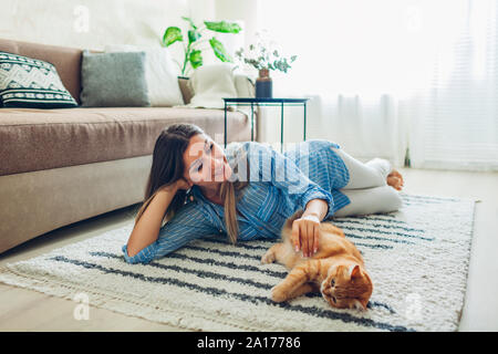 Playing with cat at home. Happy young woman lying on carpet and teasing pet. Feeling cozy speding time with ginger kitten Stock Photo