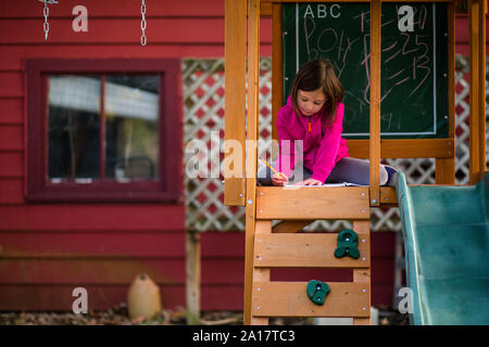 A little girl sits in a playset writing in a notebook with a pencil Stock Photo