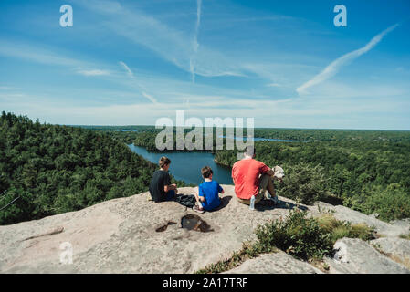 Man, boys and dog sitting on rocky summit overlooking river and trees Stock Photo