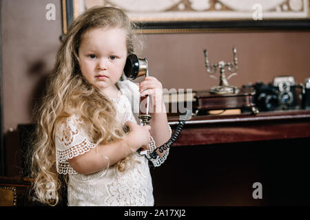 Little brooding child girl talking on a retro telephone in the room alone with sad emotions. Stock Photo