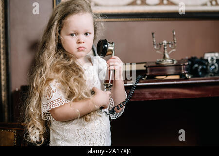 Little brooding child girl talking on a retro telephone in the room alone with sad emotions. Stock Photo