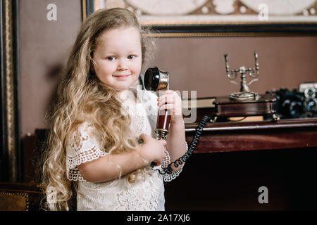 Little brooding child girl talking on a retro telephone in the room alone with sad emotions. Stock Photo