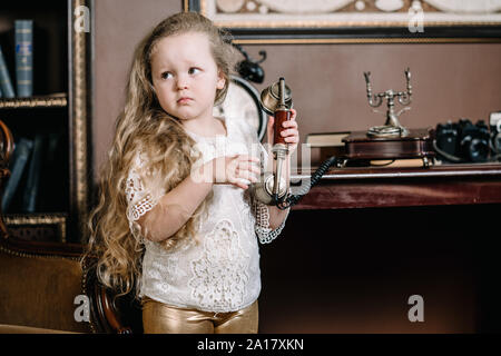 Little brooding child girl talking on a retro telephone in the room alone with sad emotions. Stock Photo