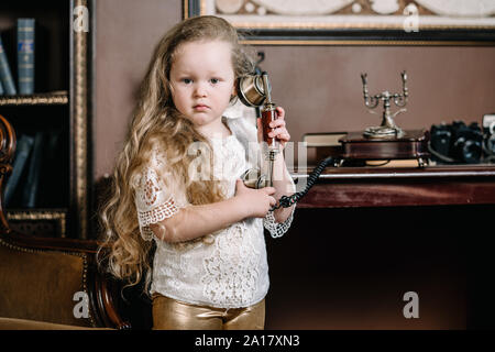 Little brooding child girl talking on a retro telephone in the room alone with sad emotions. Stock Photo