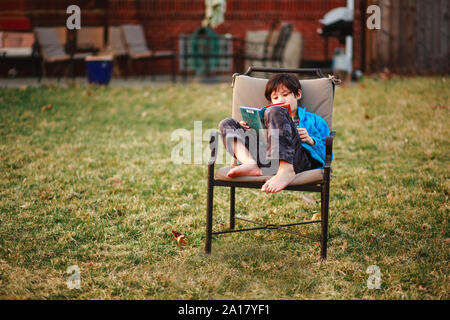 A happy boy reads a book barefoot in backyard in cool weather Stock Photo