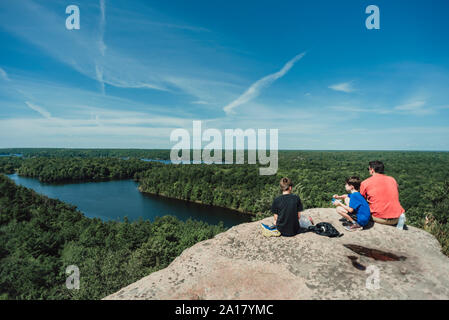 Man, boys and dog sitting on rocky summit overlooking river and trees Stock Photo