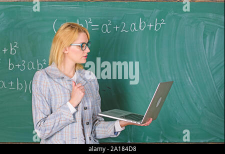 Distance education concept. Lady teacher wear eyeglasses holds laptop surfing internet. Teacher blonde woman with modern laptop surfing internet chalkboard background. School innovation. Stock Photo