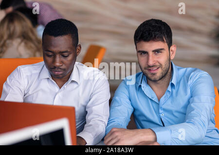 Two diverse work colleagues smiling while sitting together in an office discussing paperwork and working on a laptop Stock Photo