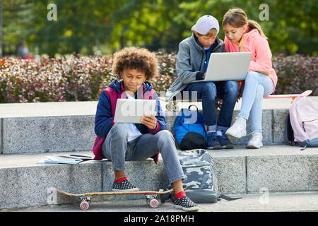 African boy using tablet pc while sitting on stairs with skateboard outdoors with his friends using laptop in the background Stock Photo