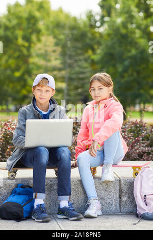 Portrait of boy and girl sitting on skateboards and looking at camera while using laptop computer outdoors in the park Stock Photo