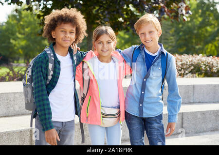 Portrait of group of friends with backpacks embracing each other and smiling at camera while standing in the park Stock Photo