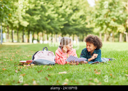 Two school children lying together on green grass with books and talking to each other during reading Stock Photo