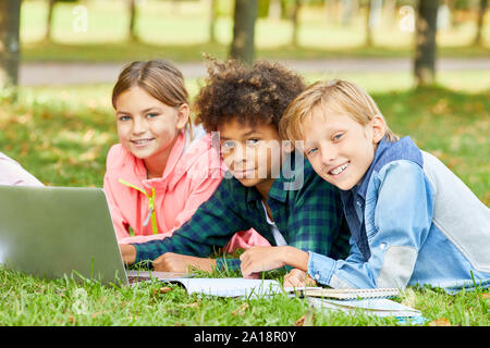 Portrait of multiethnic group of children lying on green grass and looking at camera while using laptop computer together Stock Photo