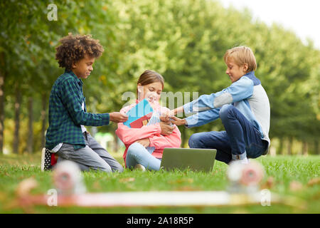 Two boys picking up the notebooks from girl while they sitting on the grass and doing homework together Stock Photo