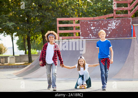 Portrait of two boys holding hands with the girl and riding her on skateboard at skateboard park outdoors Stock Photo