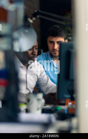 Two diverse work colleagues smiling while sitting together in an office discussing paperwork and working on a laptop Stock Photo