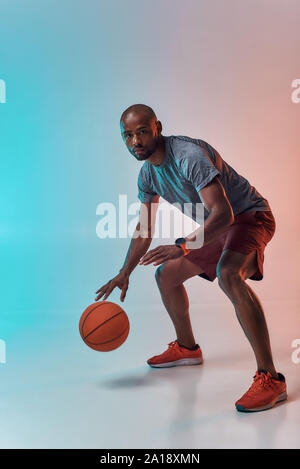 Ready to win. Full length of confident young african man in sports clothing playing basketball while standing against colorful background. Active lifestyle. Professional sport. Web banner Stock Photo