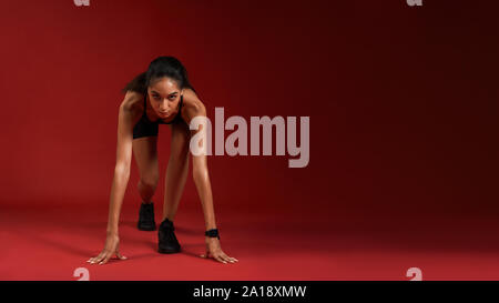 I am ready Portrait of young athlete woman in sportswear going to run while sitting isolated over red background. Fitness and work out concept. Horizontal shot Stock Photo