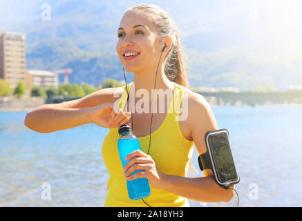 Happy cheerful female athlete with armband for smart phone open power drink bottle after daily training and looking away outdoors. Stock Photo