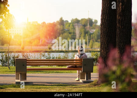 Lonely young woman sitting on a bench in the park near the lake on sunset Stock Photo