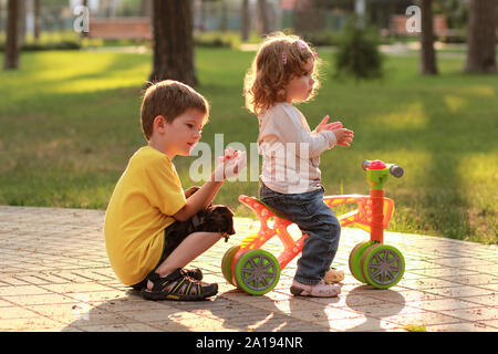 Little girl riding a small bike in the sunlit summer park and playing with a brother Stock Photo