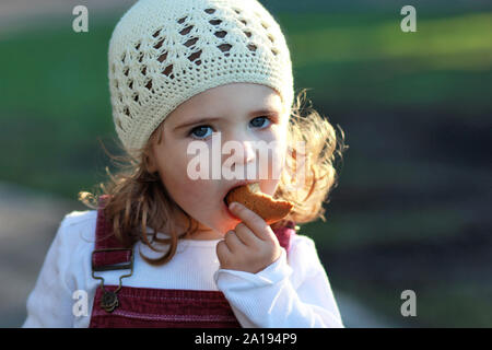 Close up portrait of cute one year old girl in a white knitted hat eating cookie on a stroll in the park, blurred background Stock Photo