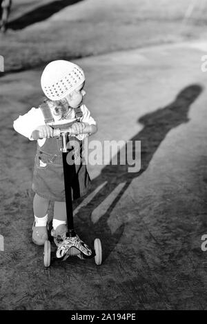 Black and white portrait of a stylish one year old girl riding a scooter and her shadow on a lovely summer day in the park Stock Photo