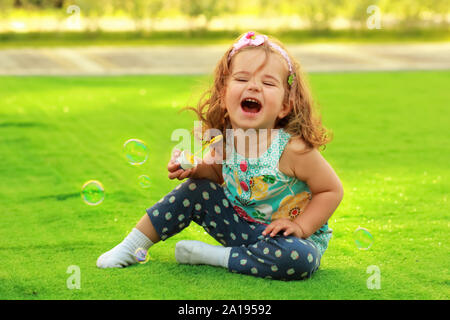Laughing one year old girl learning to blow soap bubbles and having fun sitting on the sunlit lawn Stock Photo