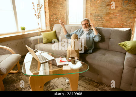 Senior man working with laptop at home - concept of home studying. Caucasian male model sitting on sofa and talking on phone or serfing in internet, watching cinema, making notes while webinar's on. Stock Photo