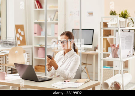 Young businesswoman in eyeglasses sitting at her office desk in front of laptop computer and using her mobile phone Stock Photo