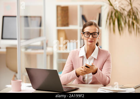 Portrait of young secretary in eyeglasses holding mobile phone and smiling at camera while working at her workplace on laptop Stock Photo