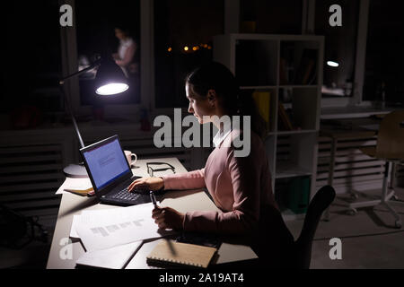 Serious manager sitting at the table working with documents and using laptop she working over project till late at office Stock Photo